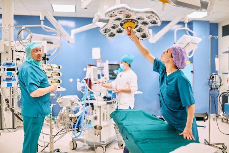Leikkausalissa kolme ammattilasta työasuissa, ei potilasta. Nainen kurottaa säätämään lamppua leikkaussängyn yllä.