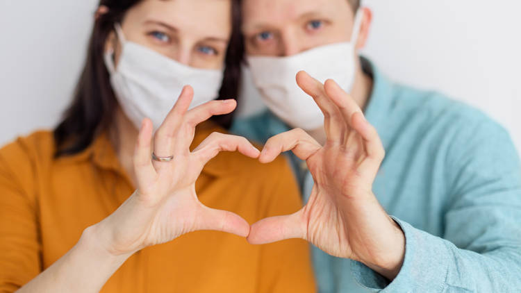Kaksi henkilöä, joilla on maskit kasvoillaan, muodostavat yhdessä sormistaan sydämen