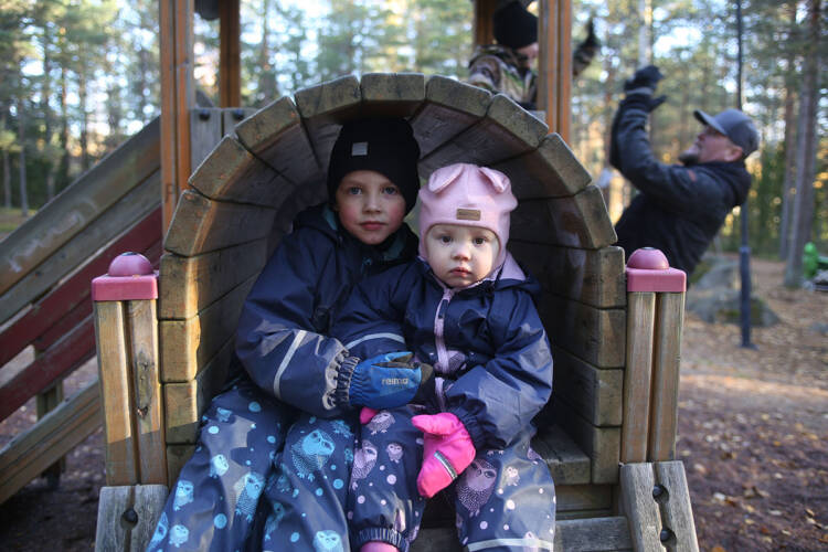 Kaksi lasta istuu kiipelytelineen tunnelissa ja katsoo kameraan. Taustalla Sami Kinnunen tekee yläfemmat lapsen kanssa. 