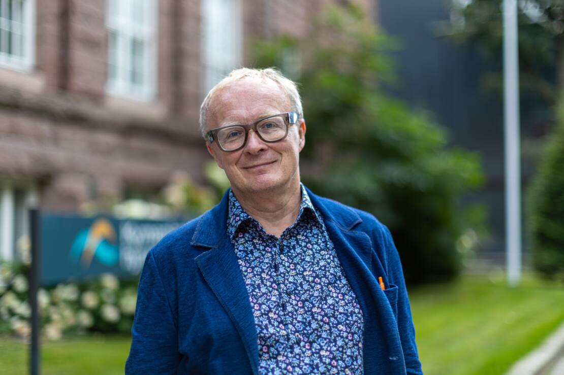 Silmälasipäinen hymyilevä mies seisoo ulkona tiilirakennuksen edessä sininen pikkutakki päällään.